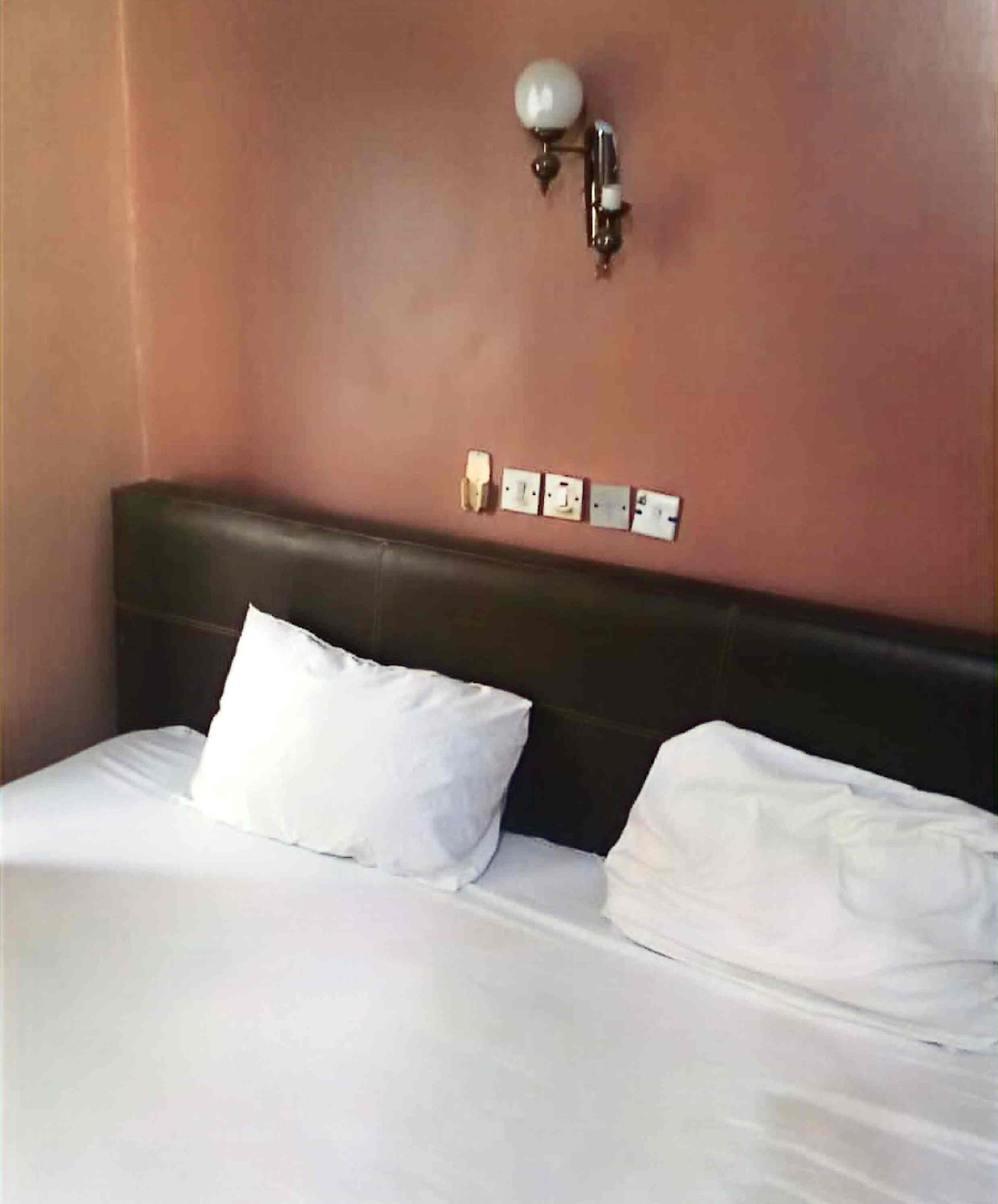 A hotel room with a single bed, white bedding, a dark headboard, and a wall-mounted light fixture.
