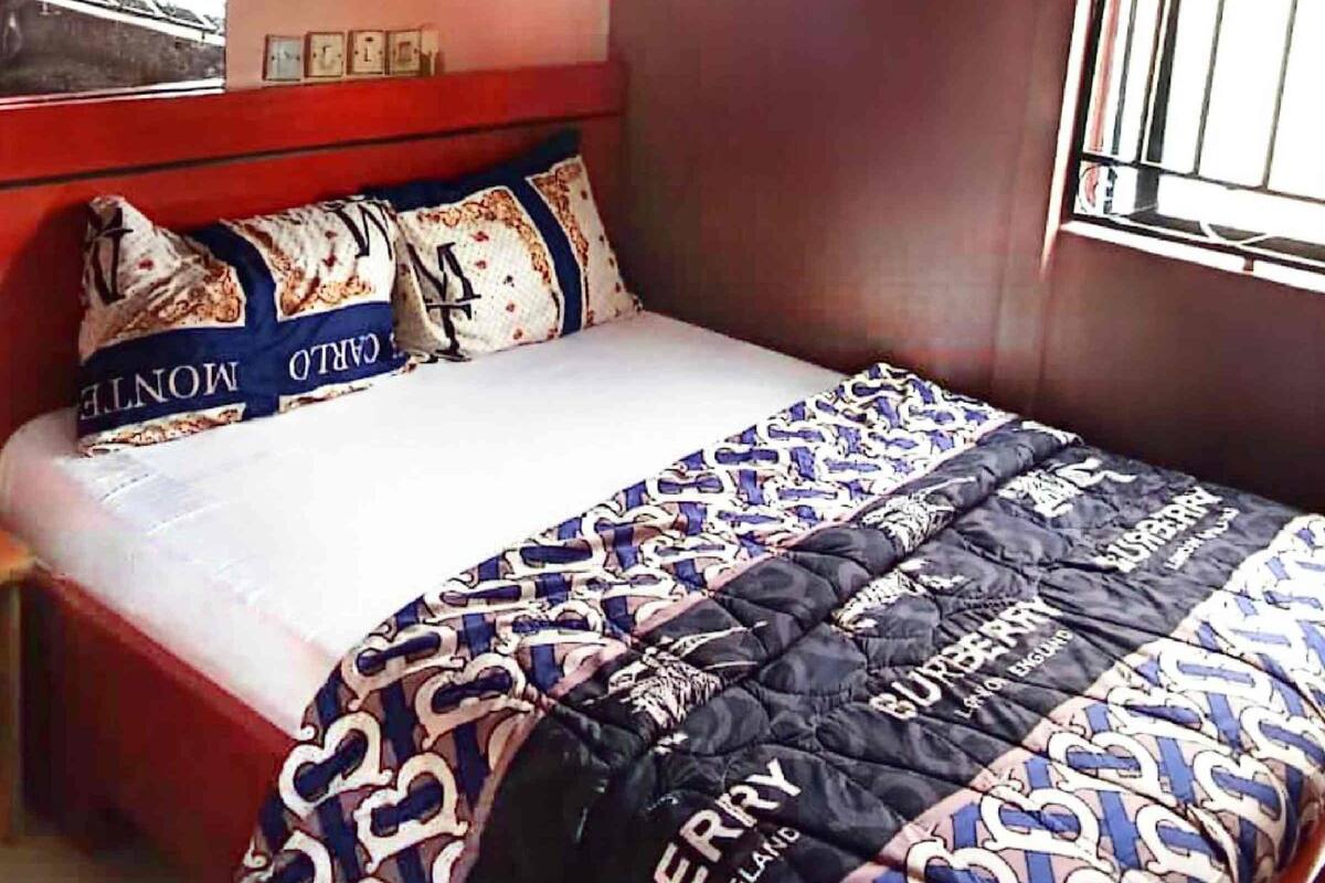 Quarto com cama, janela com cortinas, aparelho de ar condicionado e luminária fixada na parede.
