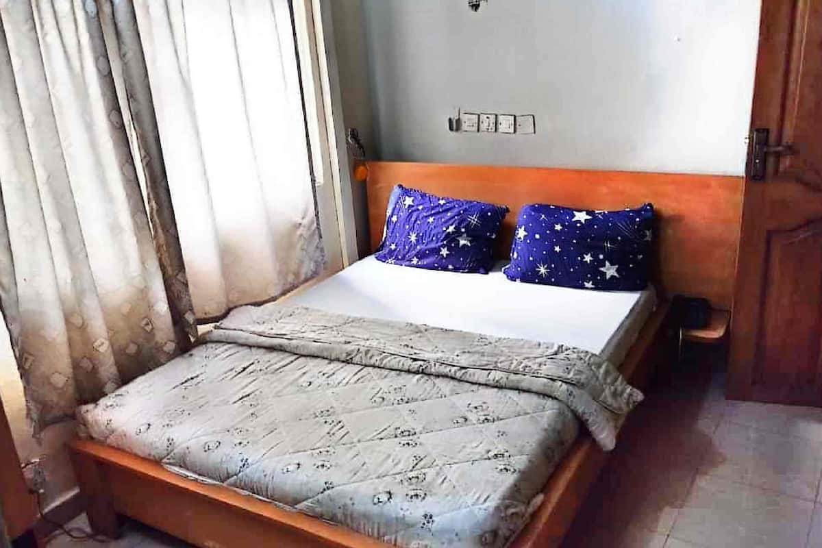 Uma cama de solteiro com colcha estampada e duas almofadas com padrão de estrelas, cabeceira de madeira, interruptor de luz e uma porta à direita.
