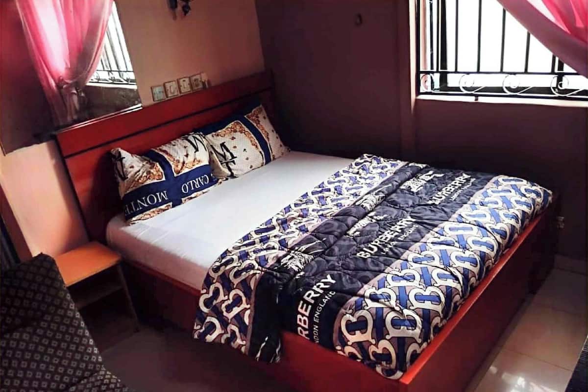 Uma cama bem arrumada com colcha estampada e almofadas, uma cabeceira de madeira, uma pequena mesa de cabeceira e uma janela com grades.