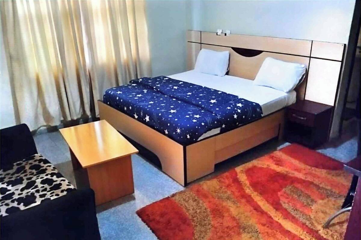 Quarto de hotel com uma cama, uma pequena mesa, uma cadeira e um tapete vermelho e laranja.