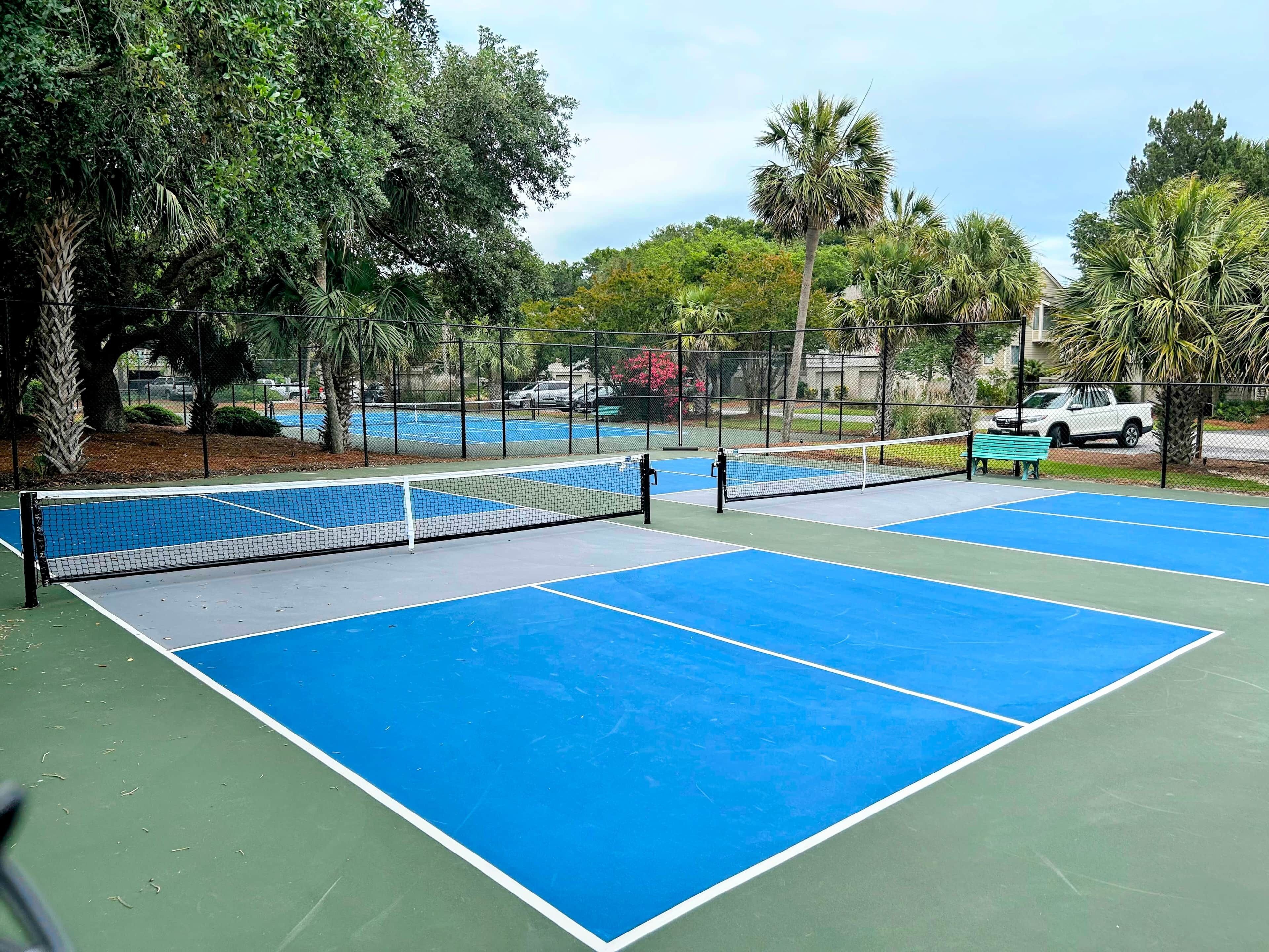 Un court de tennis en plein air, doté d’un filet, entouré d’arbres et délimité par une clôture.