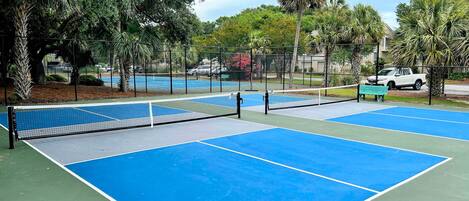 Un court de tennis en plein air, doté d’un filet, entouré d’arbres et délimité par une clôture.