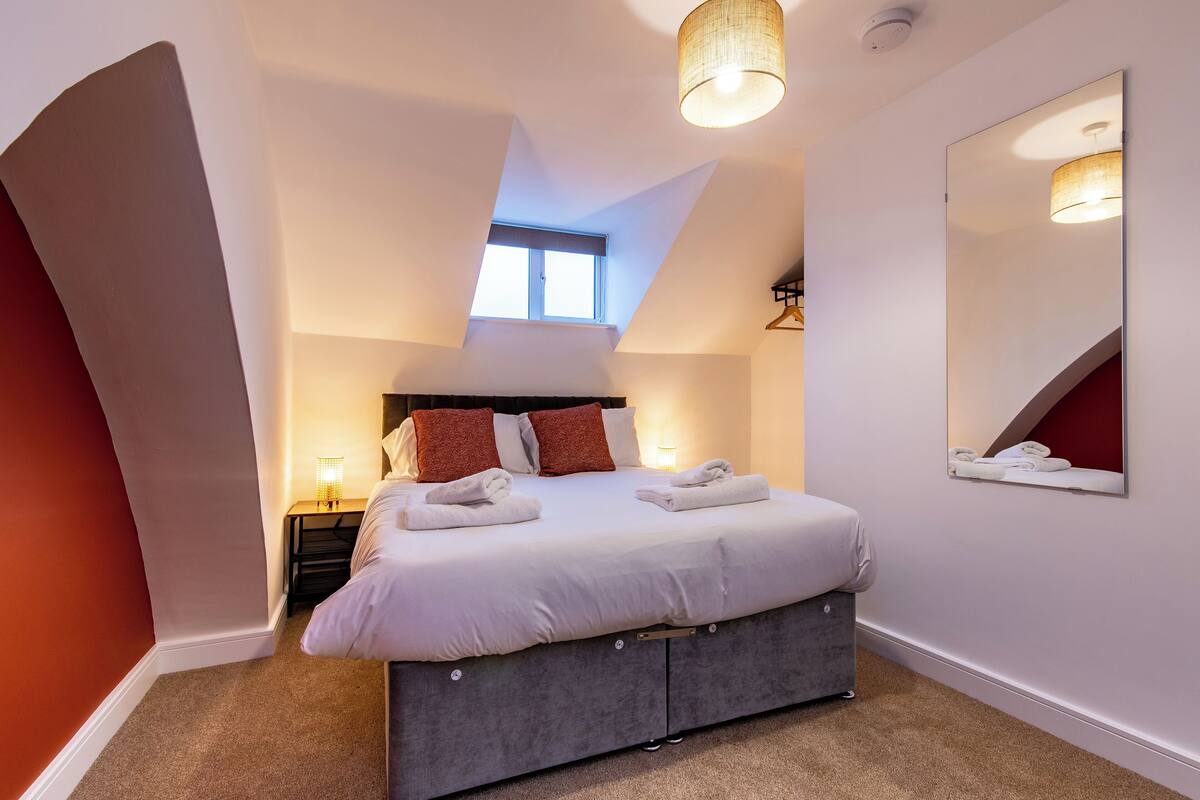 A bedroom with a bed, a bedside table, a lamp, a mirror, and a window.