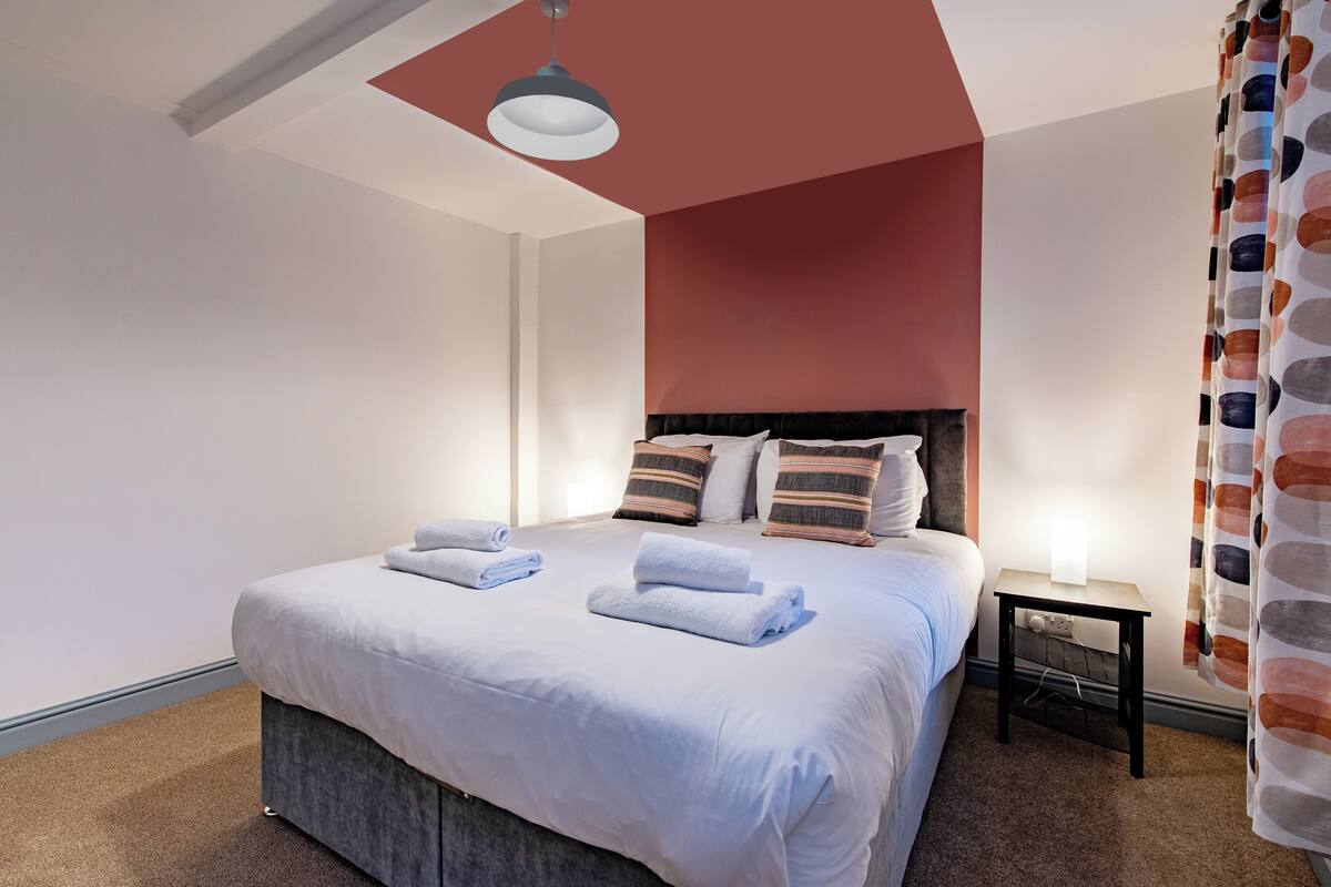 A neatly made bed with white linens and two folded towels, a bedside table with a lamp, and a patterned curtain.