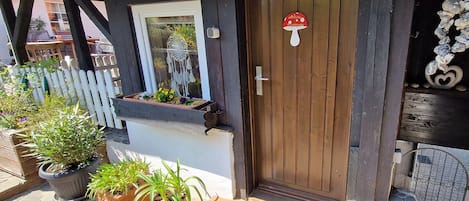 Een houten deur met een bord met daarop ‘welkom’ in de vorm van een paddenstoel, plantenpotten en een klein raam met een decoratie van een dromenvanger.