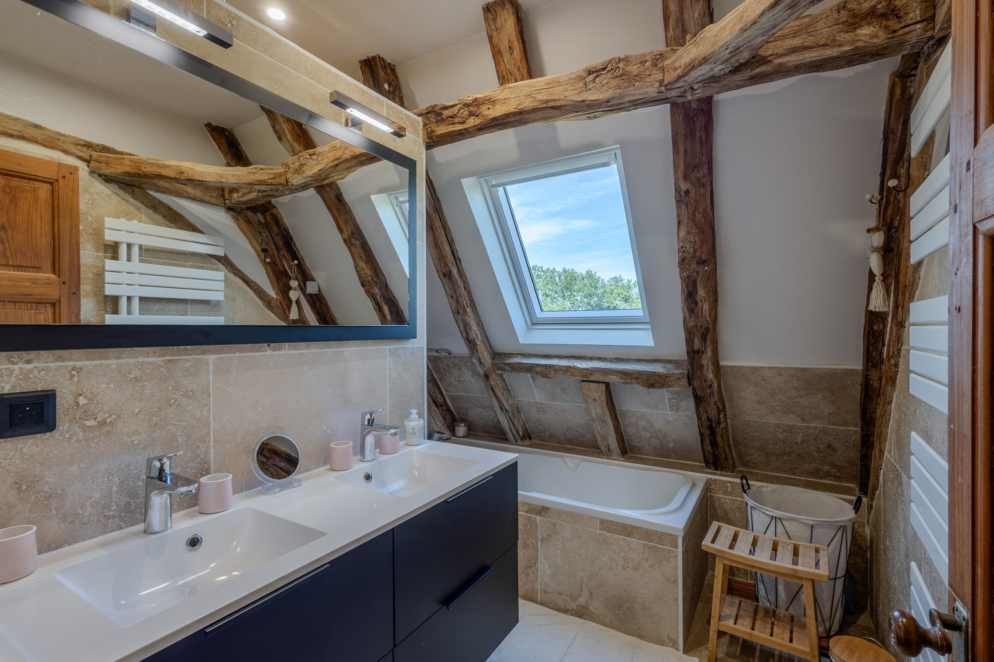 A bathroom with a large mirror, wooden beams, a skylight, a bathtub, and a wooden stool.