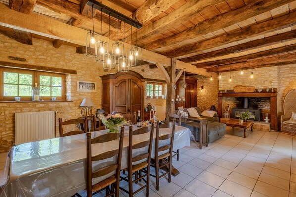 A rustic interior with exposed wooden beams, a stone wall, a dining area with a table and chairs, and a fireplace.