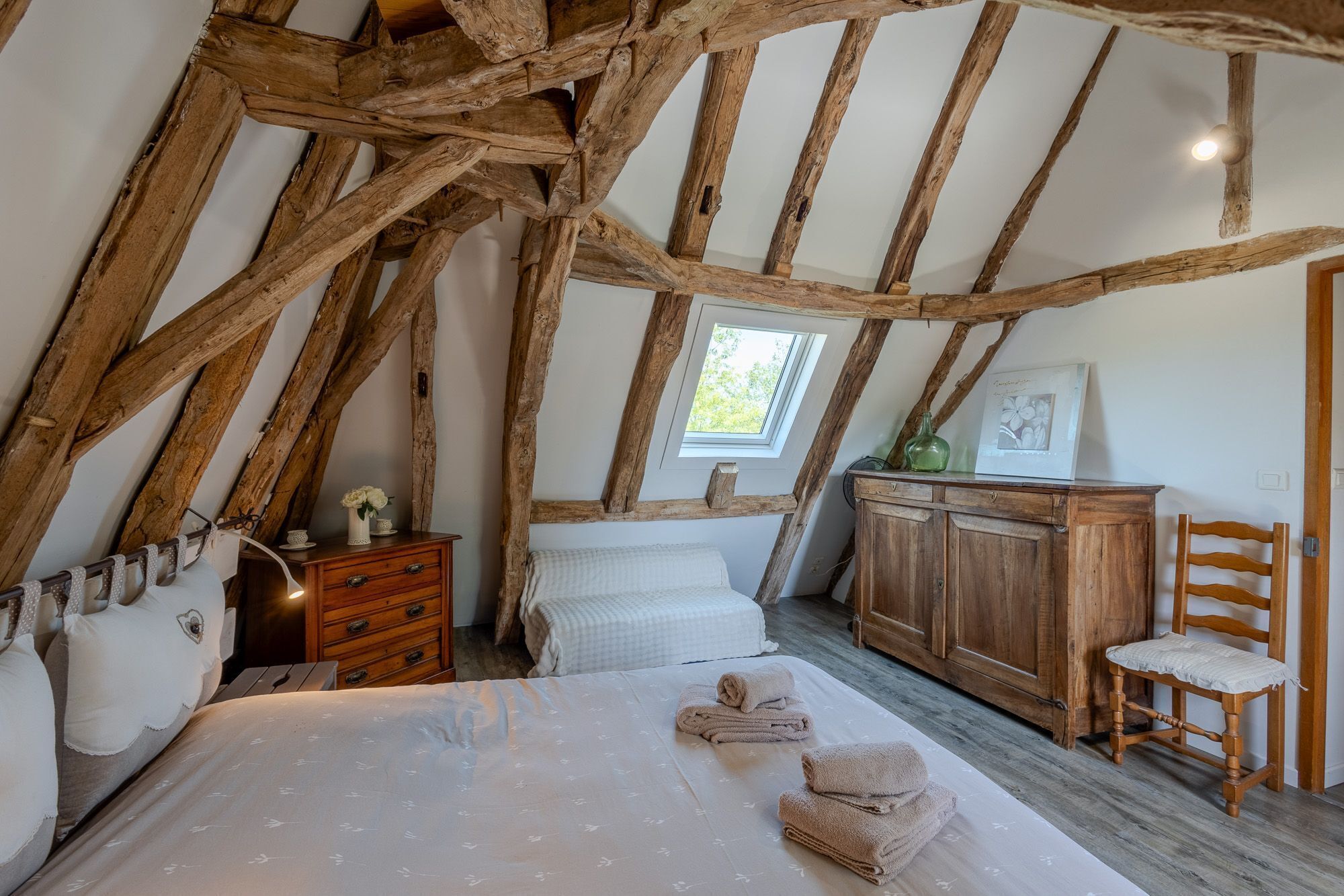 Une chambre avec des poutres apparentes, un lit avec une literie blanche, une commode, une chaise et une fenêtre donnant sur un paysage verdoyant.