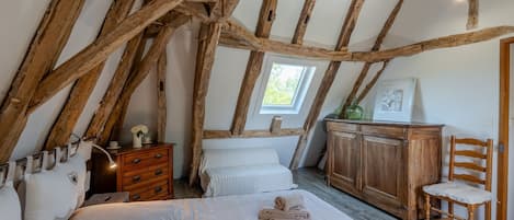 A bedroom with exposed wooden beams, a bed with white bedding, a dresser, a chair, and a window with a view of greenery.