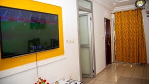 Ένα δωμάτιο με μια τηλεόραση τοποθετημένη σε έναν κίτρινο τοίχο, μια πόρτα που οδηγεί σε ένα άλλο δωμάτιο και μια κουρτίνα με σχέδια.