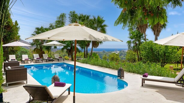 A pool area with lounge chairs and umbrellas, surrounded by greenery and a view of the ocean.