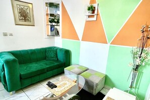 Een groene bank, een glazen bijzettafeltje met afstandsbedieningen, en een wanddecoratie met een geometrisch patroon.