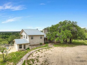 Uma casa de pedra de dois andares com telhado de metal, cercada por um caminho de cascalho e uma pequena área de jardim.