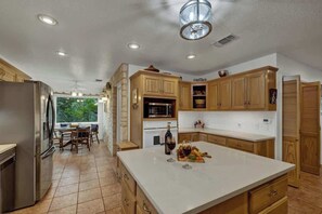 A modern kitchen with wooden cabinets, a large island, and a dining area with a view of trees. - Firewheel Serene Creek Front Home Games+Grill (Fredericksburg)