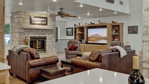 A spacious living room with leather sofas, a stone fireplace, a flat-screen TV, and a ceiling fan.