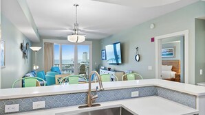 Una cocina moderna con un lavamanos de acero inoxidable, una encimera blanca y un salpicadero con diseño azul y blanco. Hay una zona de comedor con sillas verdes y vistas a la ciudad a través de amplios ventanales.