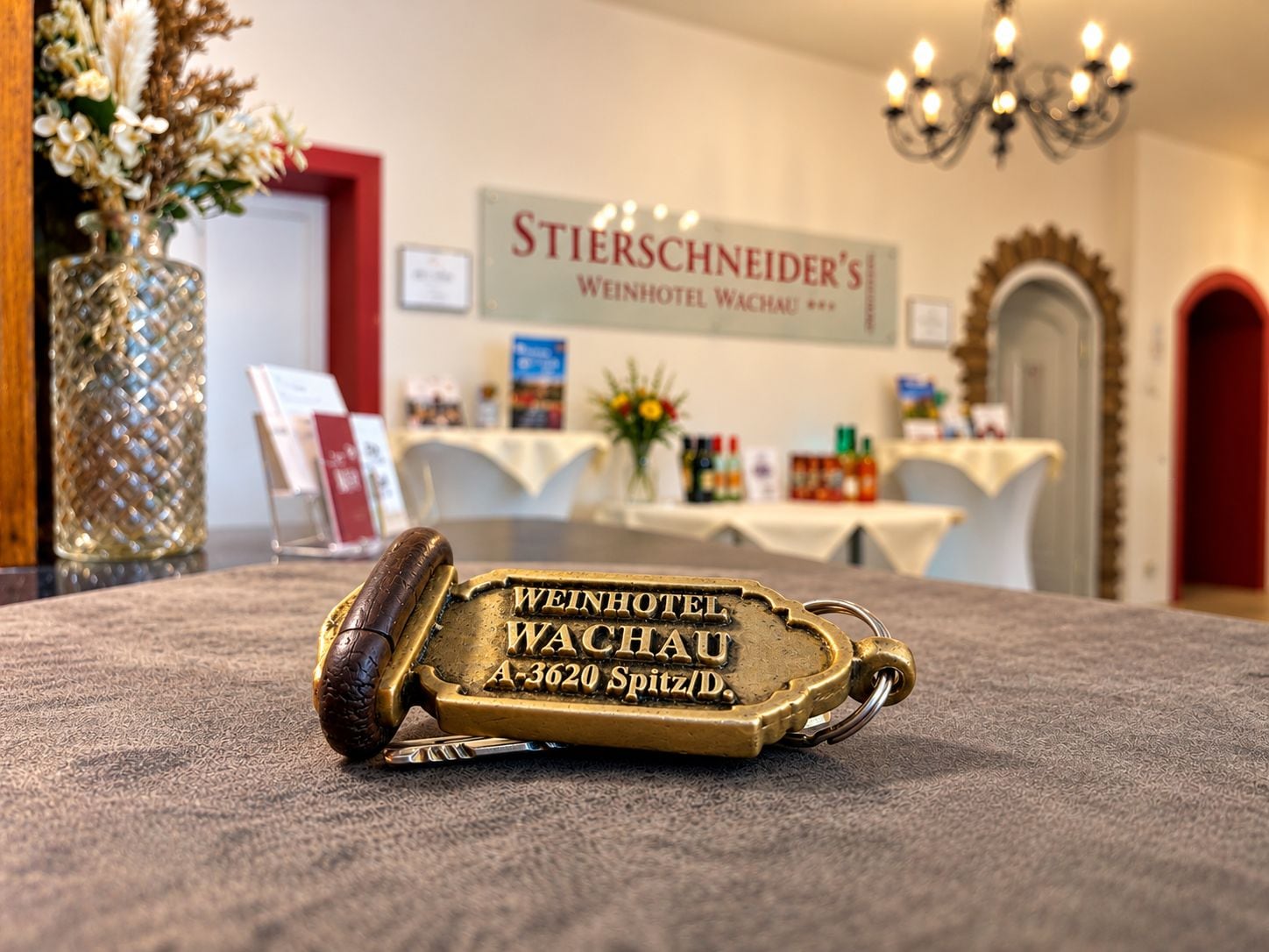 Ein mit 'Weinhotel Wachau' gravierter Messing-Schlüssel liegt auf einem Teppichboden in einer Hotelhalle mit einer Kristallleuchter und einem Schild mit der Aufschrift 'Stierschneider's Weinhotel Wachau'.