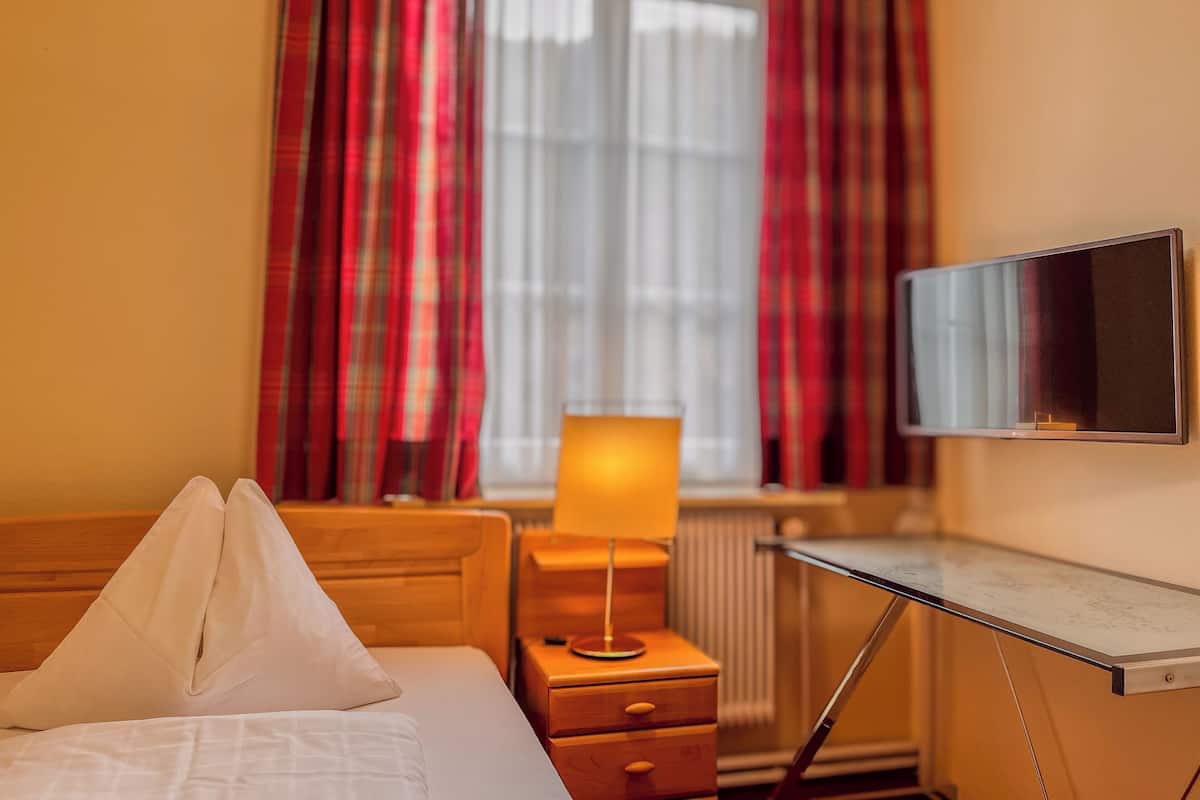 Ein Hotelzimmer mit Bett, Nachttischlampe, Schreibtisch mit Bildschirm und Fenster mit Vorhängen.