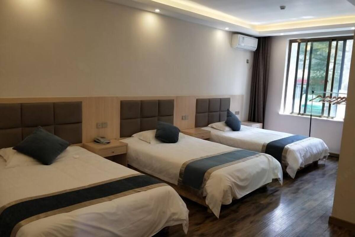 Quarto de hotel com três camas, cabeceiras de madeira, uma janela com persianas e ar condicionado instalado no teto.