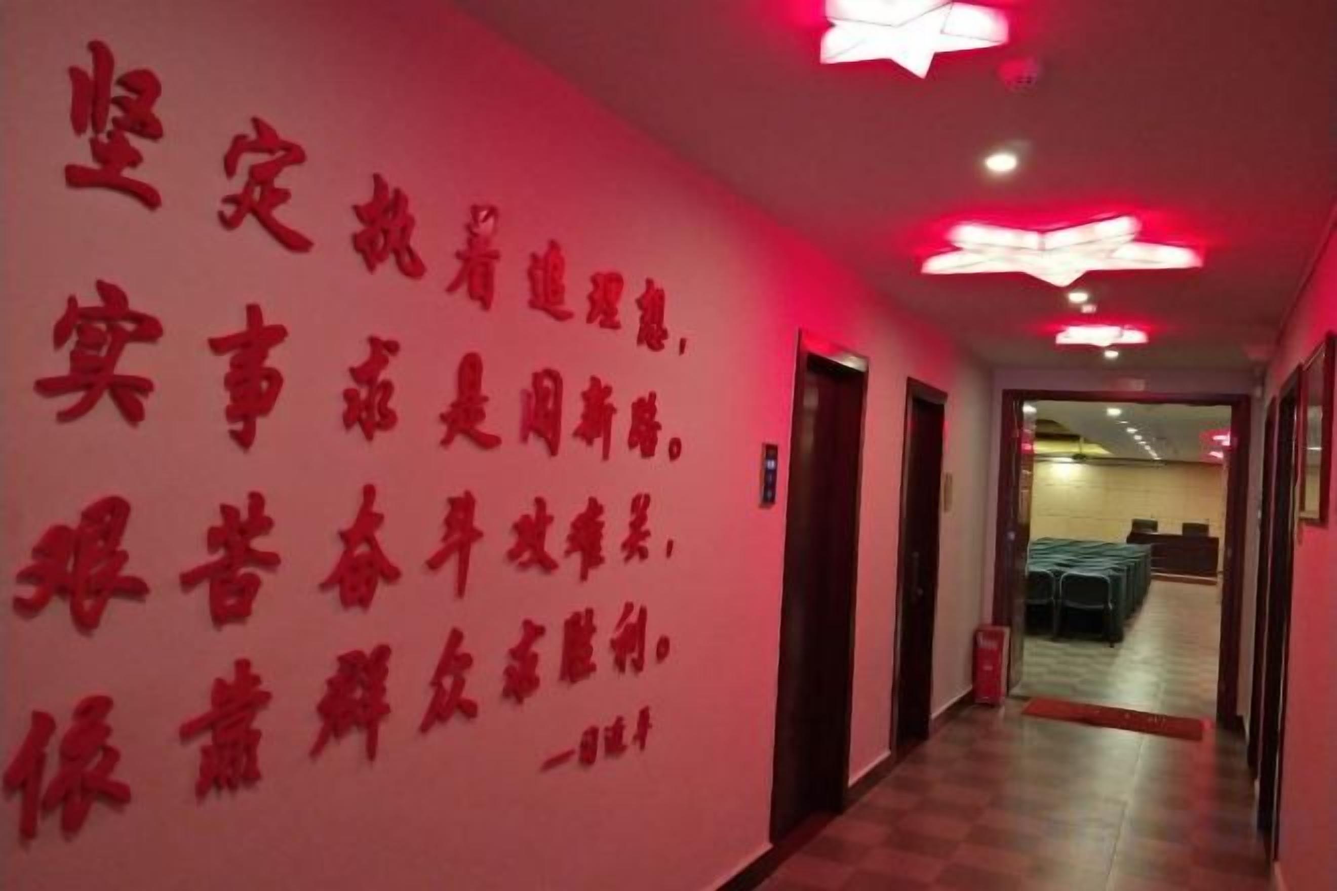 Um corredor com parede vermelha e série de texto em vermelho. Há portas à direita e um espaço com mesas e cadeiras ao fundo.