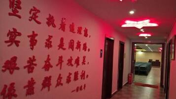 Um corredor com parede vermelha e série de texto em vermelho. Há portas à direita e um espaço com mesas e cadeiras ao fundo.