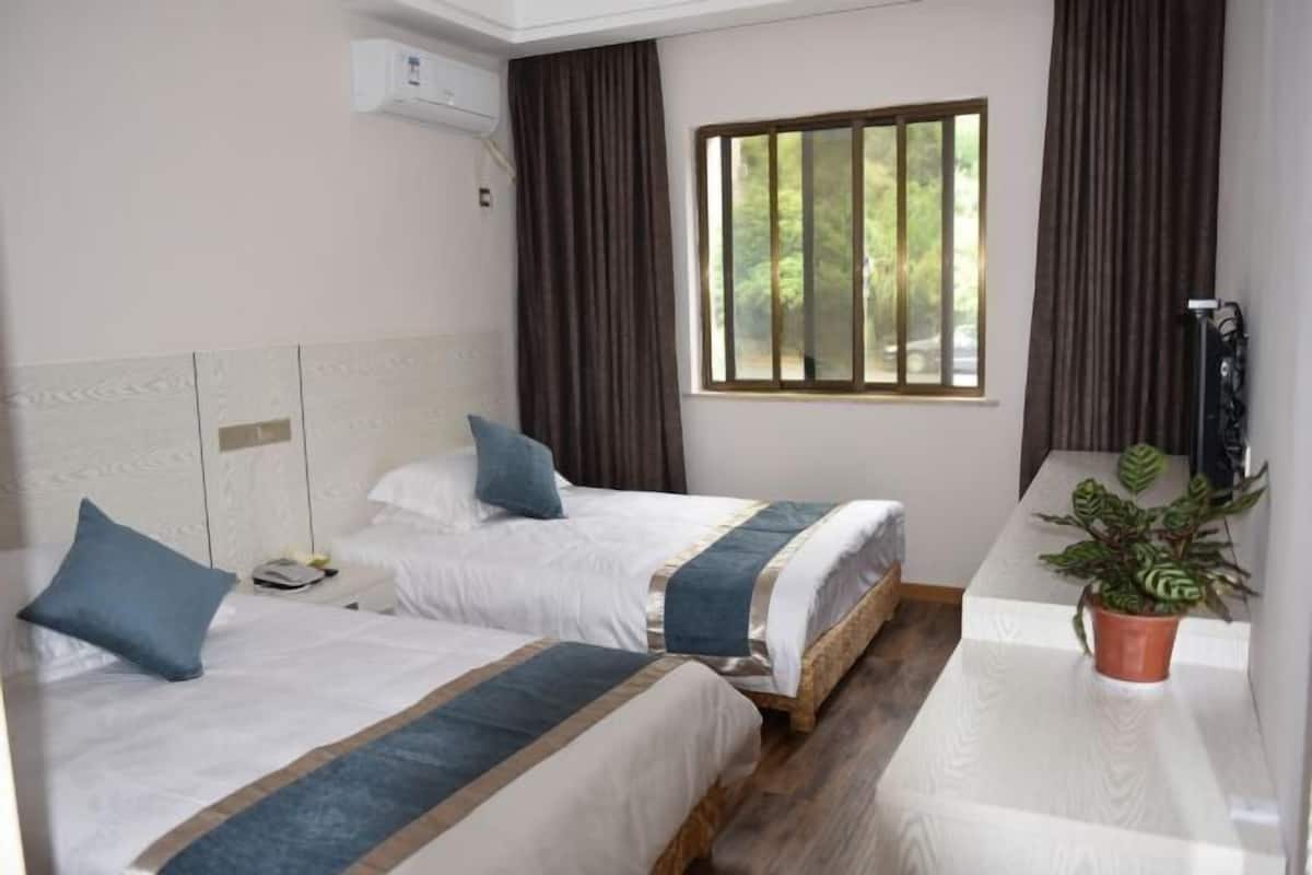 Quarto de hotel com duas camas, janela com cortinas, planta em vaso e ar-condicionado fixado na parede branco.