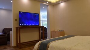 Camera d'hotel con una grande TV a schermo piatto su un supporto in legno, un letto con copriletto blu e bianco, un piccolo armadietto in legno e una finestra che lascia entrare luce naturale.