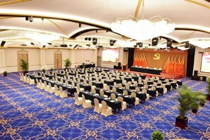 Een congreszaal met rijen tafels en stoelen, een podium met een rode achtergrond en een grote kroonluchter.