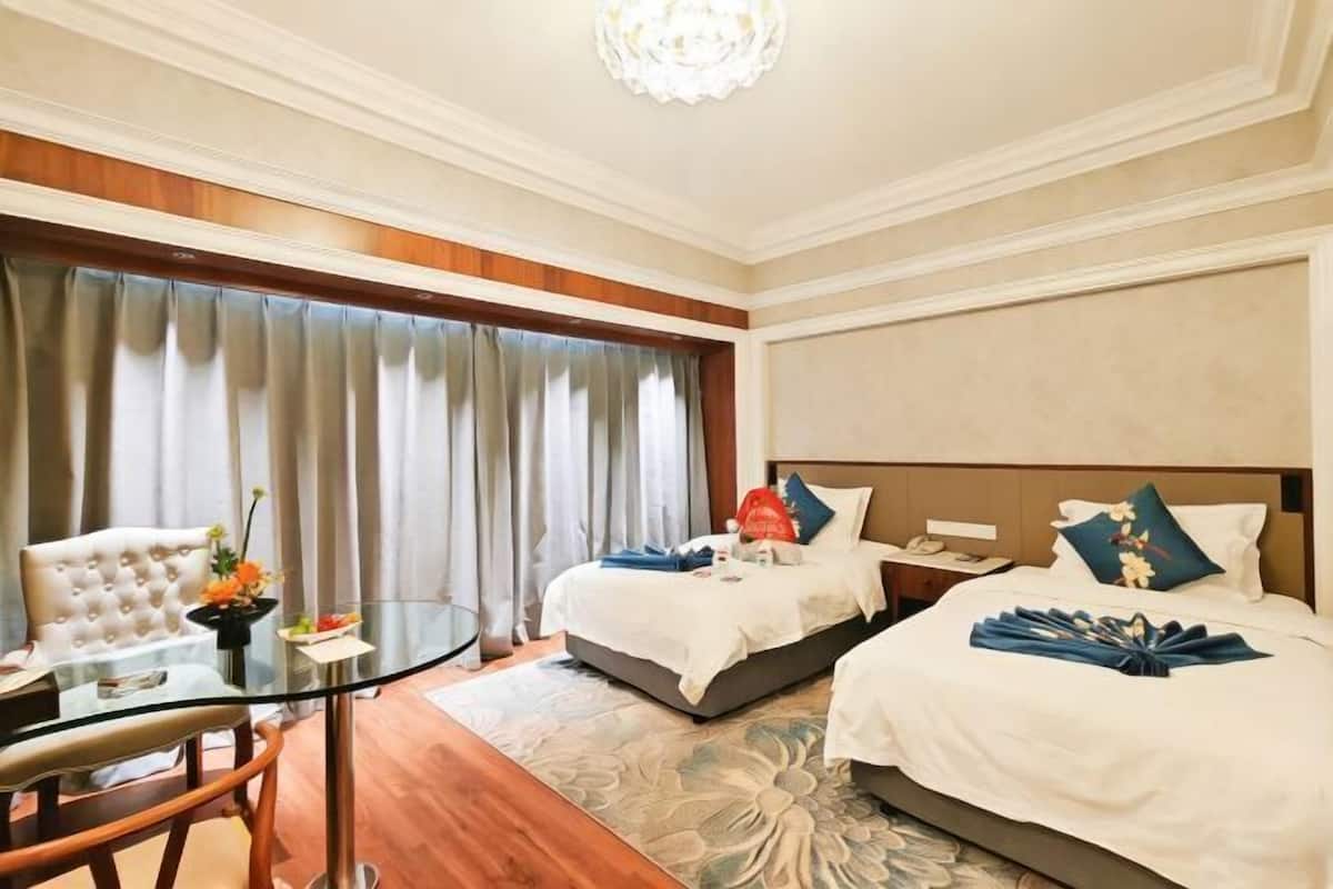 A hotel room with two beds, a dining table, a chair, and a large window with curtains.