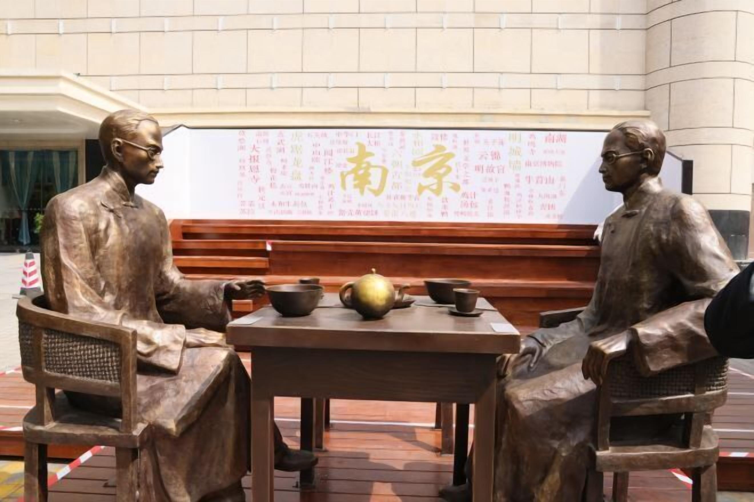 Een standbeeld van twee figuren zittend aan een tafel met een traditionele Chinese theeset, voor een whiteboard met Chinese karakters.