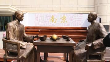Een standbeeld van twee figuren zittend aan een tafel met een traditionele Chinese theeset, voor een whiteboard met Chinese karakters.