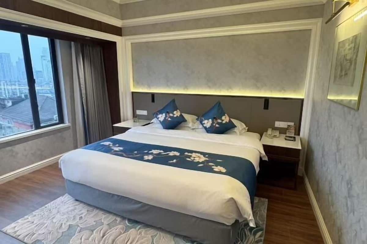 A hotel room with a large bed, a nightstand, a bedside table, a window with curtains, and a view of the cityscape.