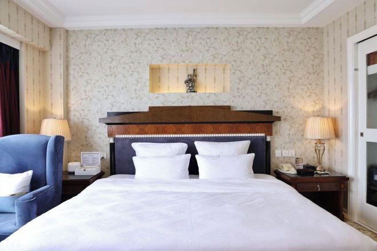 Een hotelkamer met een groot bed, twee nachtkastjes, een blauwe fauteuil en een decoratief hoofdeinde.