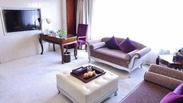 A living room with a sofa, ottoman, and armchair, a television, a desk with a lamp, and a window with curtains.