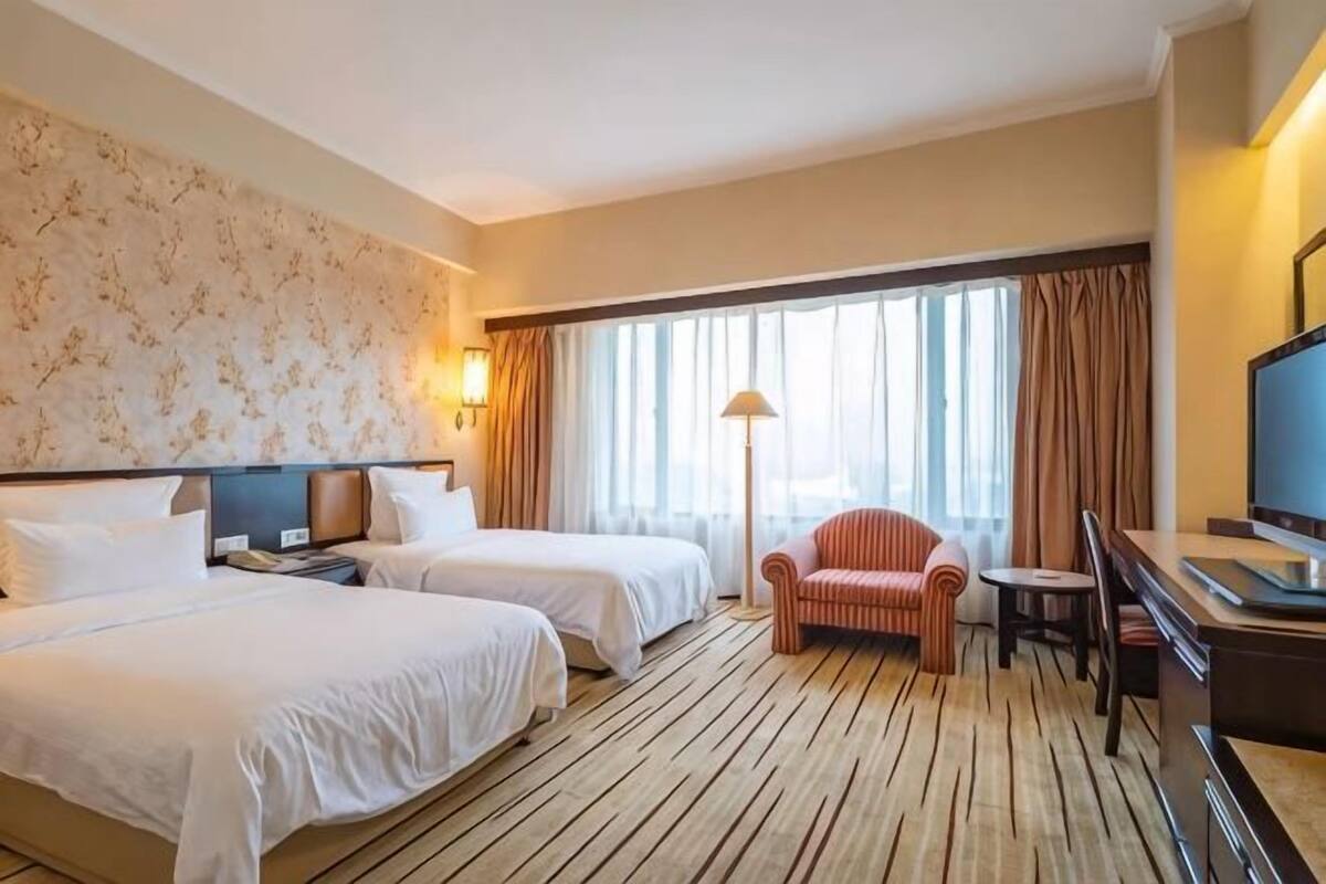 A hotel room with two beds, a desk, a chair, a TV, and a large window with curtains.