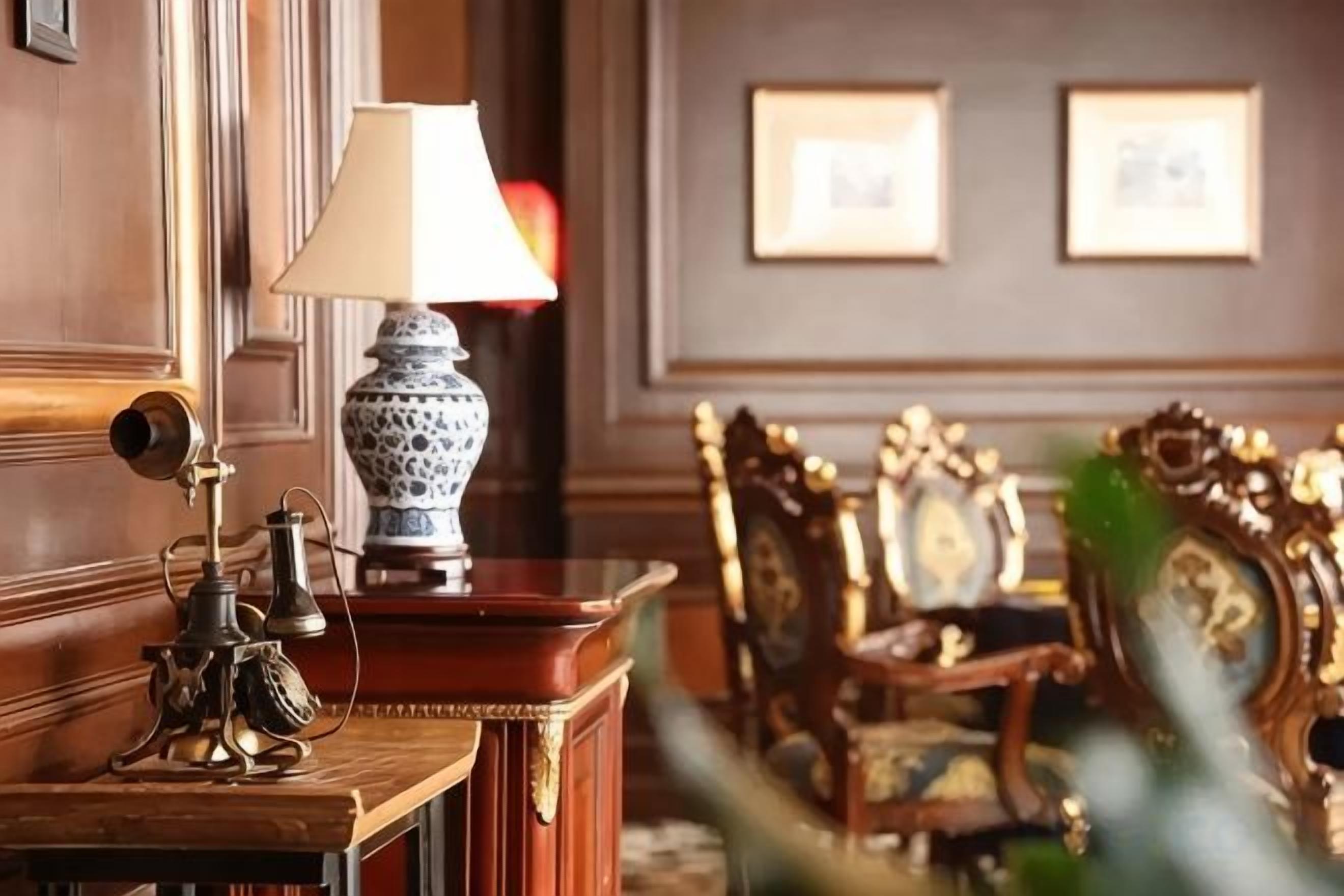 A wooden table with a lamp and a decorative object, a chair with ornate design, and framed pictures on the wall.