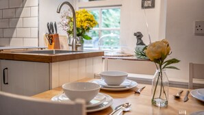 Una cucina con mobili bianchi, un piano di lavoro in legno, un lavello e una finestra che si affaccia sul verde. Si vedono ciotole, un vaso con fiori e un cartello appeso al muro.