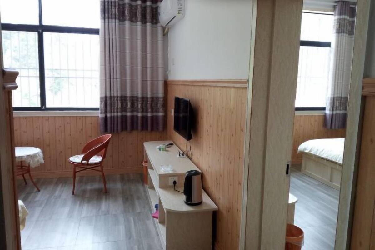 Ein Zimmer mit Holzboden, einem Einzelbett, einem kleinen Tisch mit Stuhl, einem an der Wand befestigten Fernseher und einem Fenster mit Vorhängen.