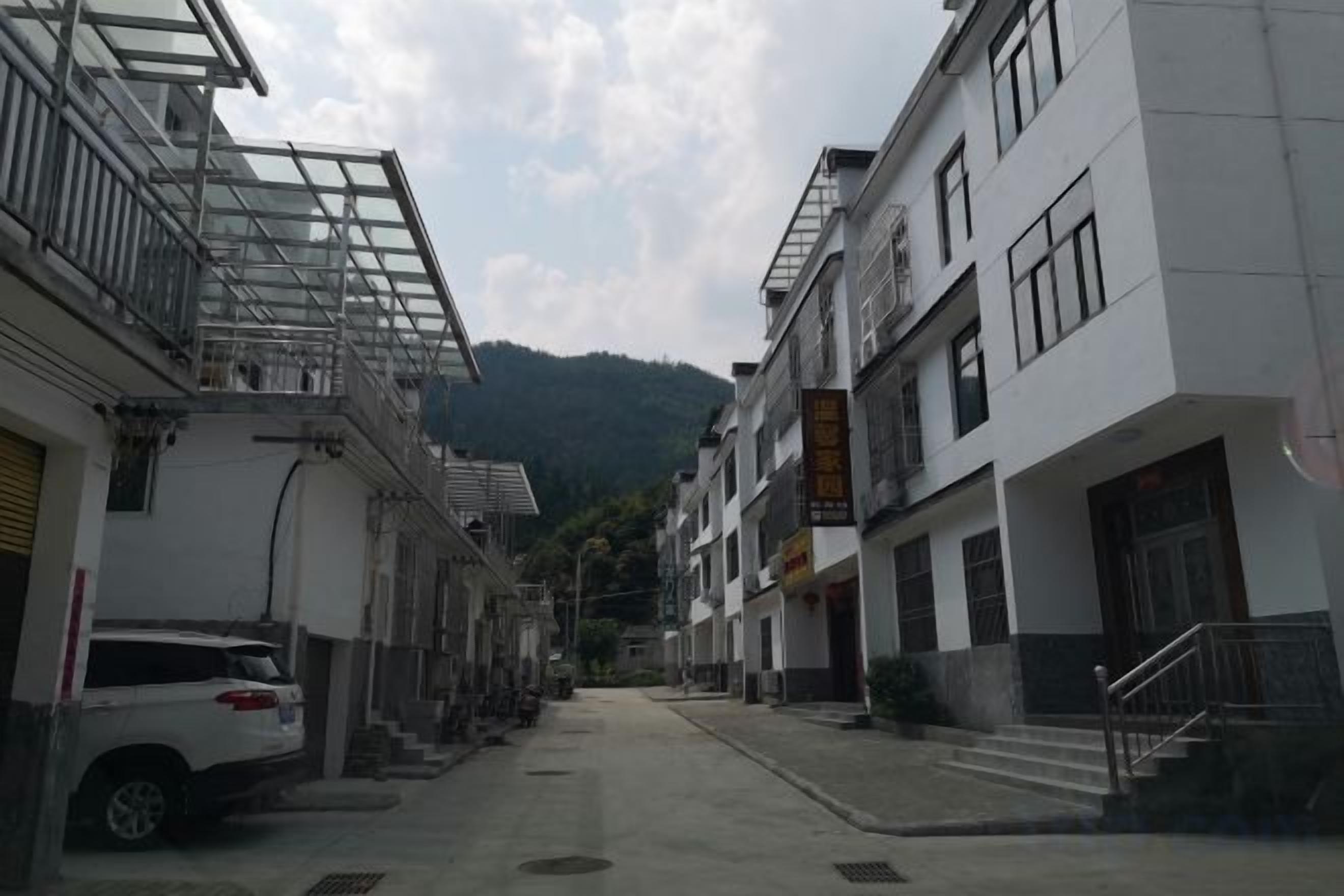 A narrow street with buildings on either side, a parked car, and a mountain in the background.