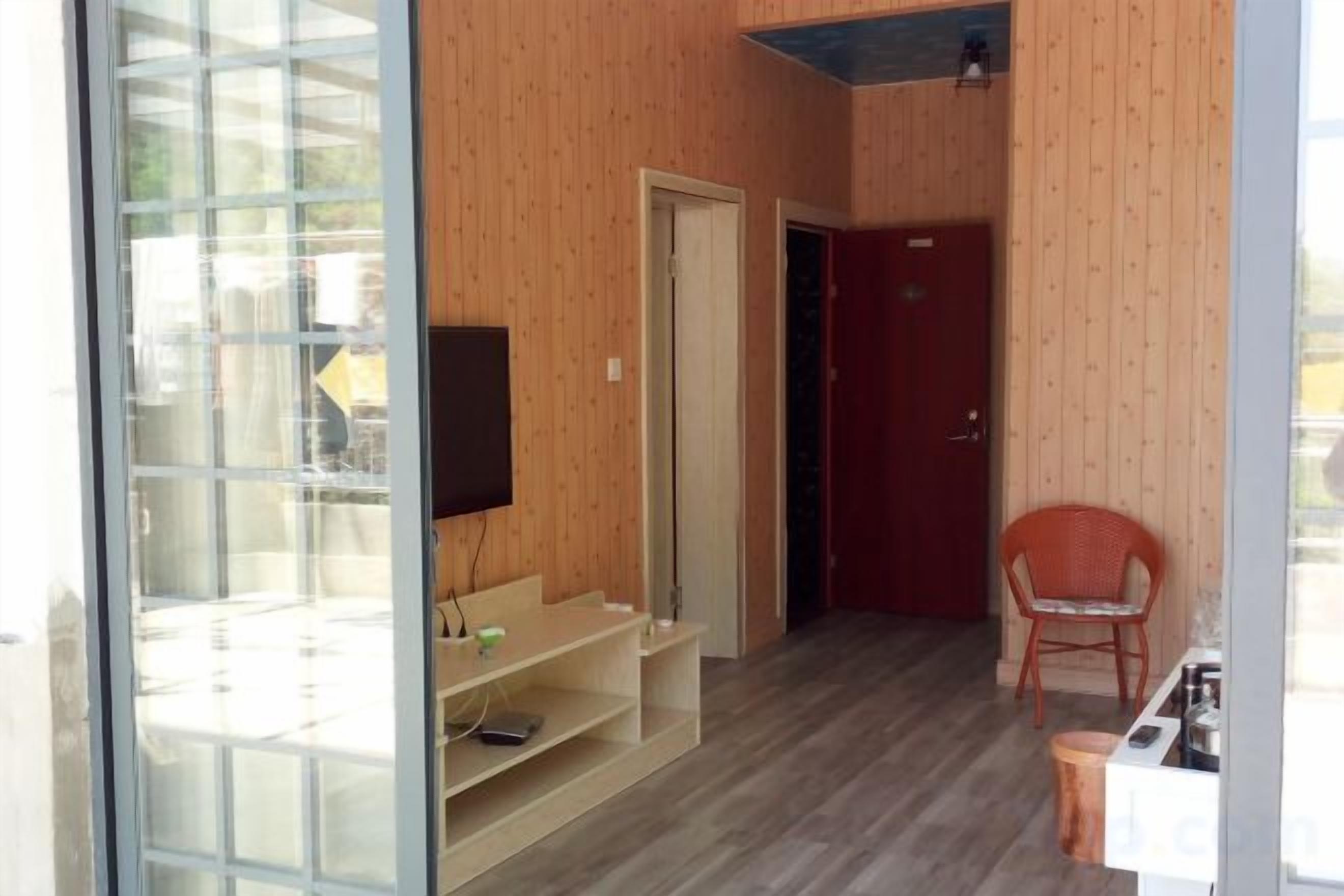 A room with a wooden floor, a TV mounted on the wall, a red door, and a chair.