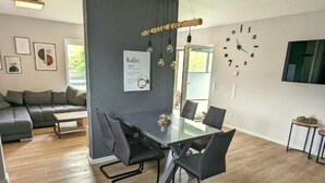 Un salon moderne avec un coin repas, comprenant un canapé gris, une table à manger avec un plateau en verre et des chaises, une horloge murale et un menu encadré accroché au mur.