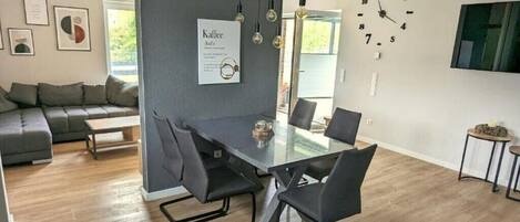 Een moderne woonkamer met een eethoek, met een grijze bank, een eettafel met glazen blad en stoelen, een wandklok en een menukaart in een lijst aan de muur.