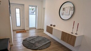 Un couloir doté d’un plancher en bois, d’un tapis rond, d’une console blanche avec un miroir et d’une porte avec un panneau de verre.