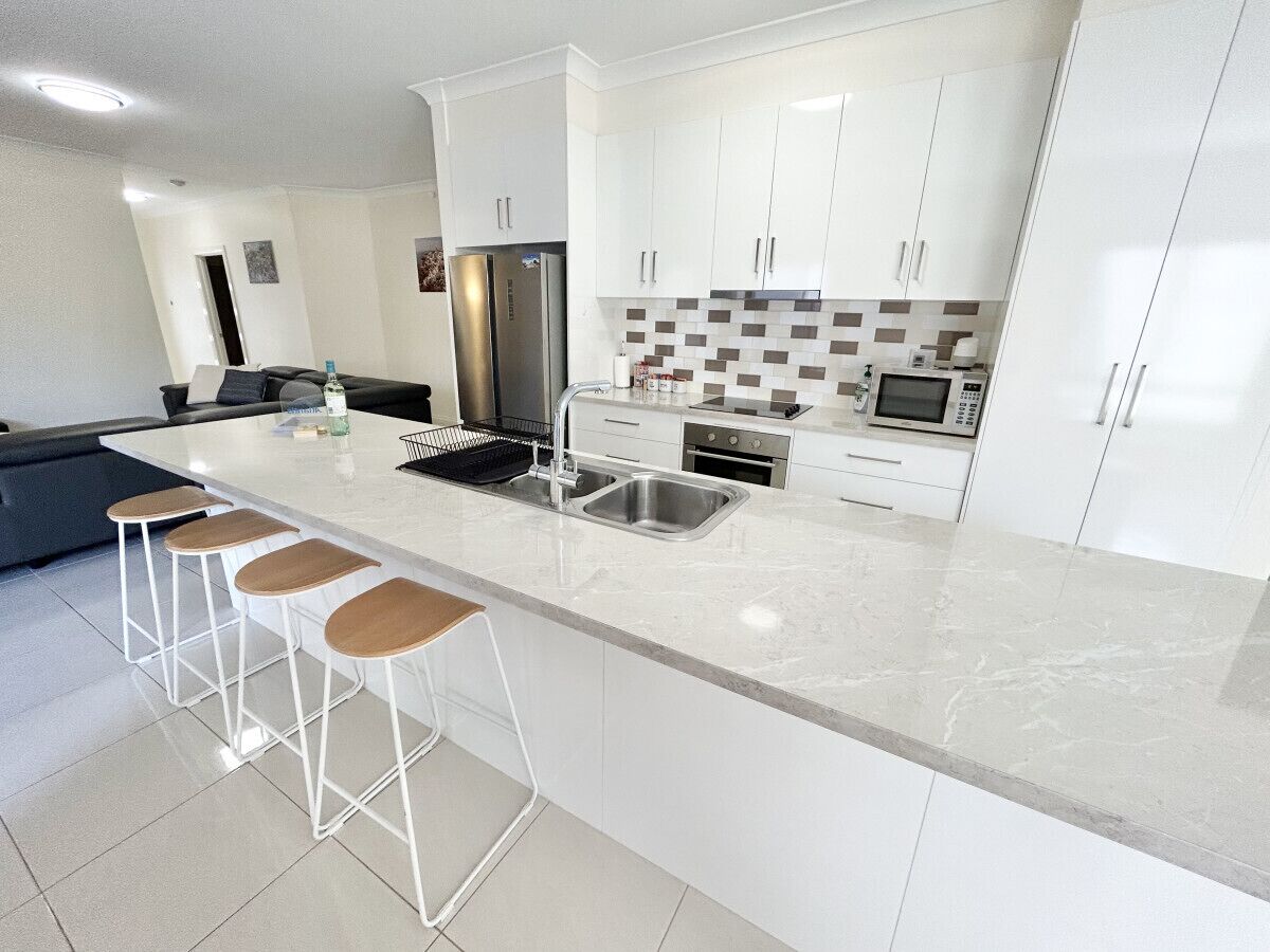 A modern kitchen with white cabinets, a marble countertop, a stainless steel refrigerator, a microwave, and a sink. There are three wooden stools at the island.