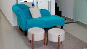 A teal sofa with a white pillow and two round ottomans with wooden legs in a modern room with a tiled floor. - Pousada Praia By Bia (Penha)