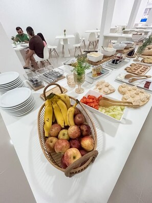 A buffet setup with various fruits, pastries, and a dining area with white chairs and tables. - Pousada Praia By Bia (Penha)
