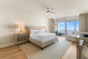 A spacious bedroom with a large bed, a ceiling fan, a nightstand with lamps, a sofa, and a view of the outdoors through large windows. - Turquoise C2208 (Orange Beach)