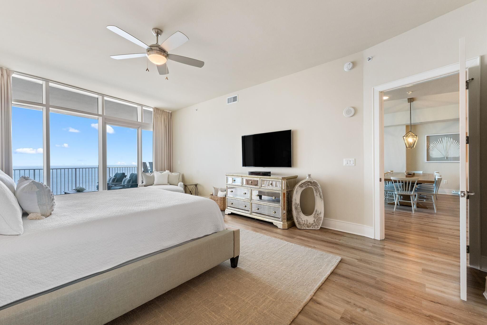 Un dormitorio espacioso con una cama grande, ventilador de techo, televisión y vistas al océano a través de amplias ventanas.