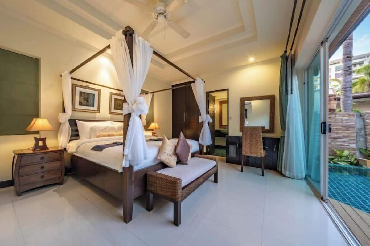 A four-poster bed with a canopy, a wooden nightstand, a bench, and a large window with a view of a pool and greenery.